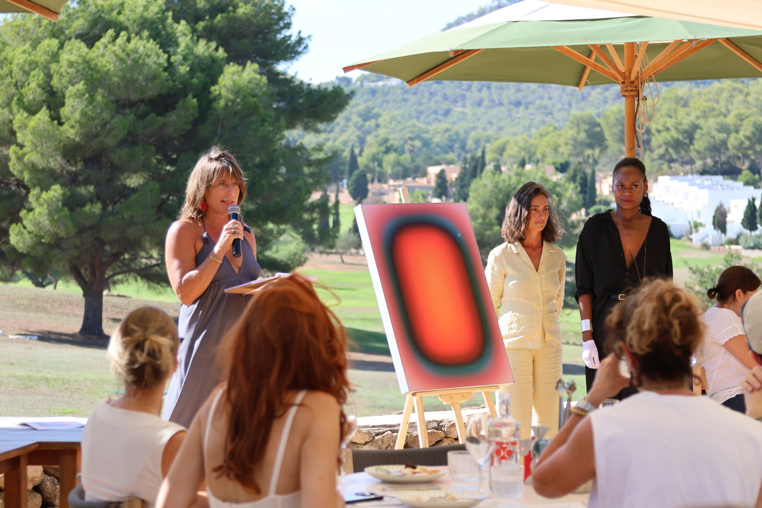 An auctioneer speaks beside a painting on an easel during an open-air Art Initiatives benefit lunch in Ibiza.