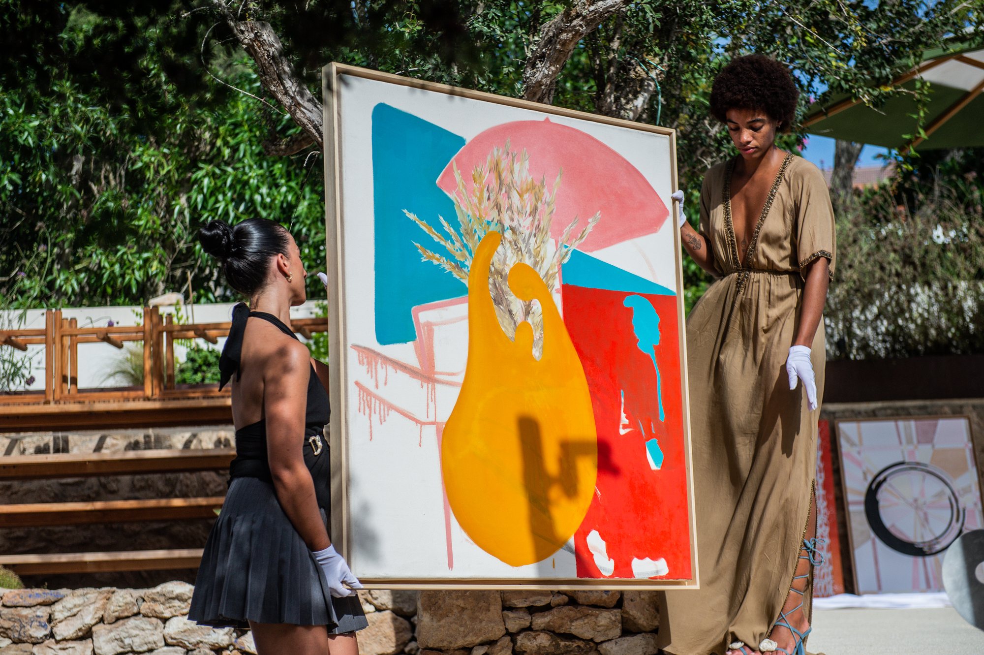 Two presenters in white gloves show a large colourful painting to guests at an open-air Art Initiatives benefit auction in Ibiza.