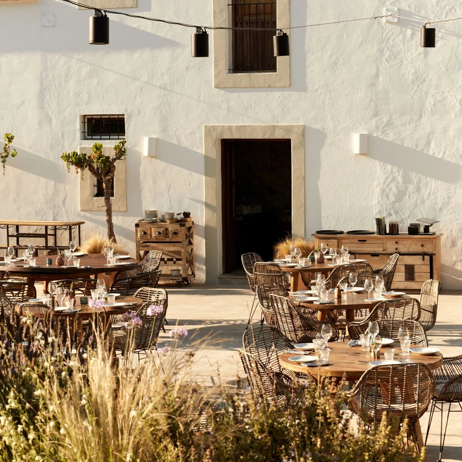 La terraza del patio de Juntos House en Sant Mateu d&rsquo;Albarca: mesas preparadas para la comida ante una casa ibicenca tradicional blanca, con hierbas silvestres y guirnaldas de luces.