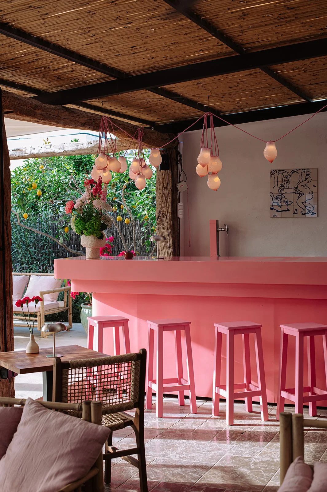 La barra rosa de Mira Cocktail Lounge en Sant Miquel, con taburetes rosas, lámparas de cristal y flores, y la terraza del jardín al fondo.