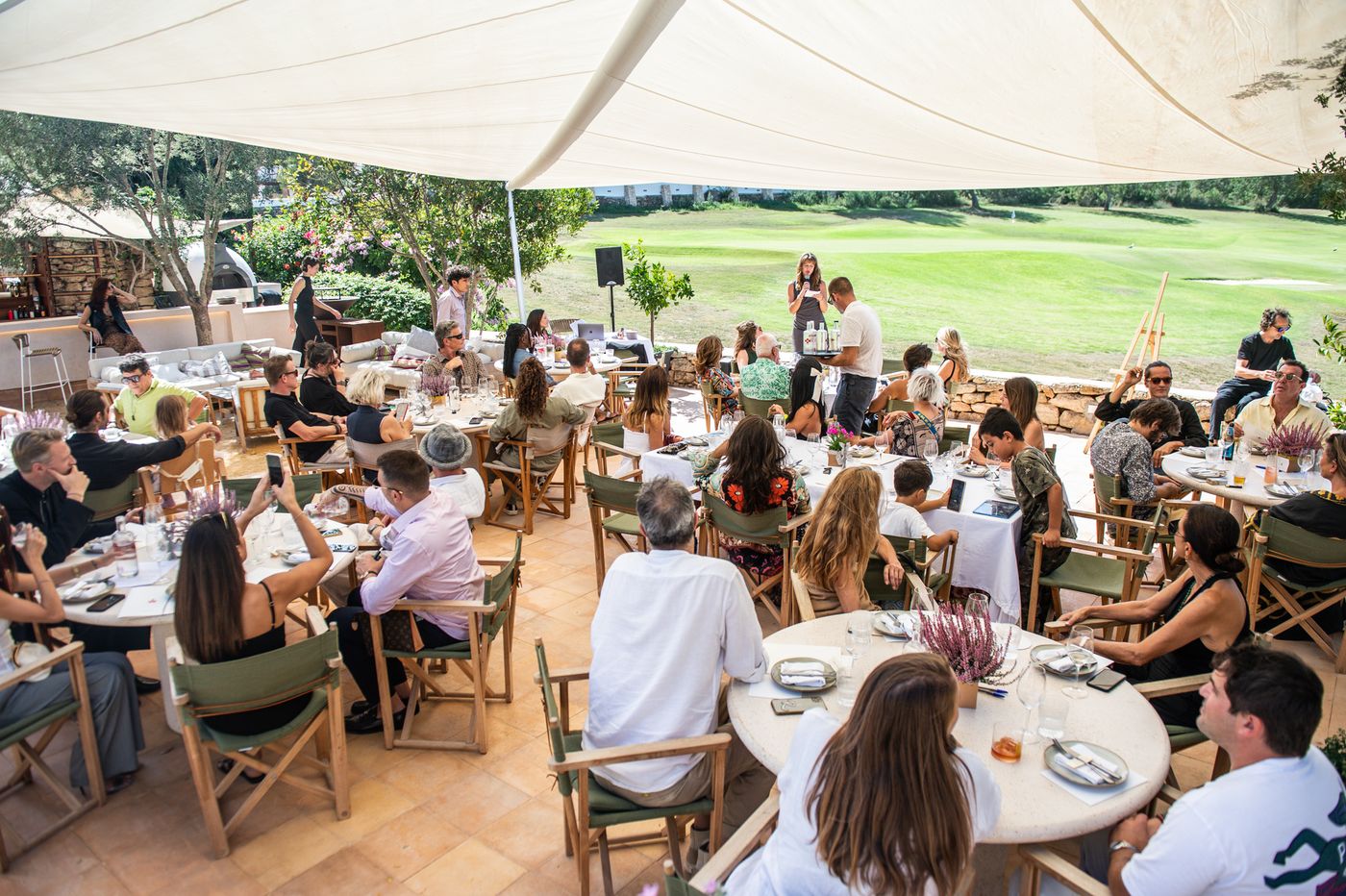 A wide view of the benefit lunch: guests at round tables under a shade sail, green hills beyond.