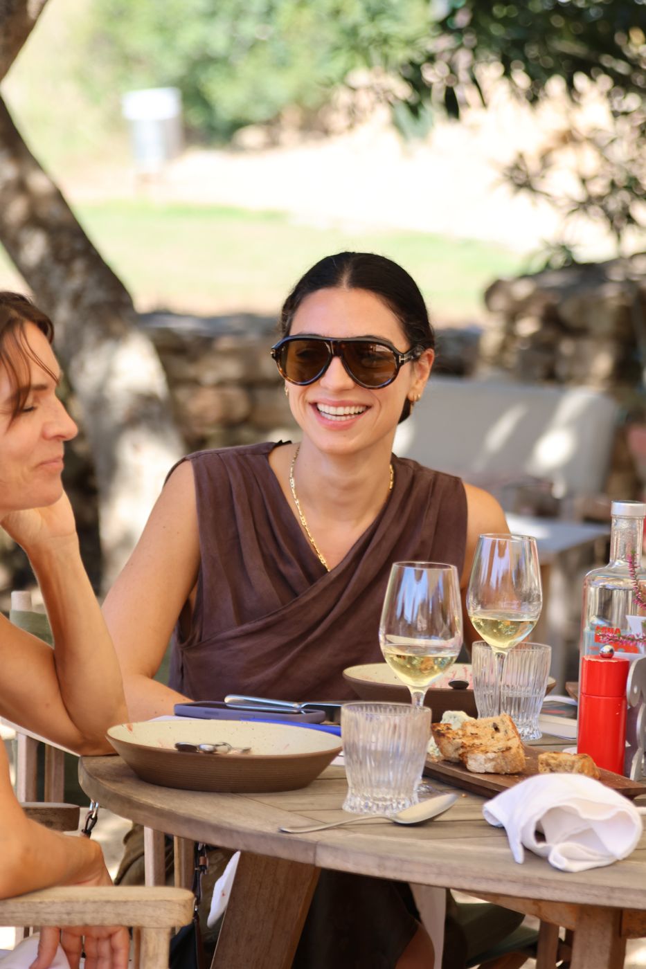 A guest in sunglasses smiles at a lunch table with a glass of white wine.